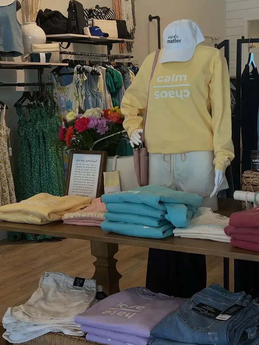Clothing boutique display with folded sweaters, hanging dresses, and a yellow sweatshirt mannequin