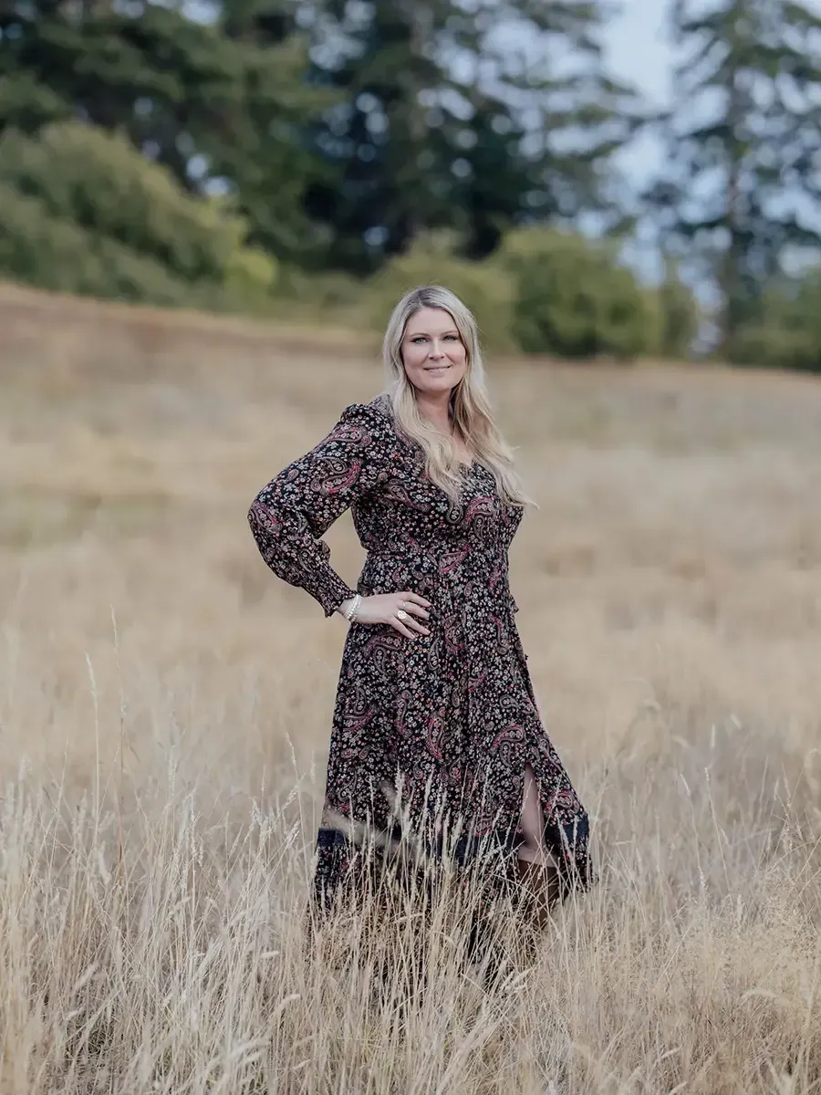 Woman standing in a grassy field, smiling with one hand on her hip, wearing a dark floral dress.