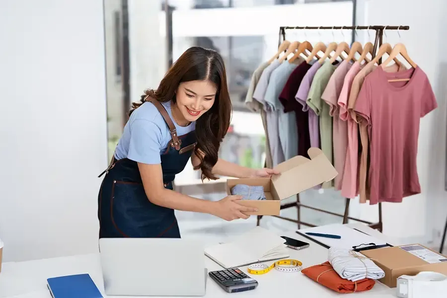 Woman packing a box at a clothing shop counter beside a rack of pastel dresses
