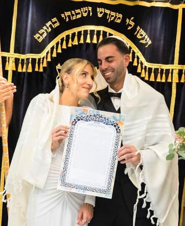 Newlyweds hold marriage certificate, smiling.  Ceremony background with Hebrew script.