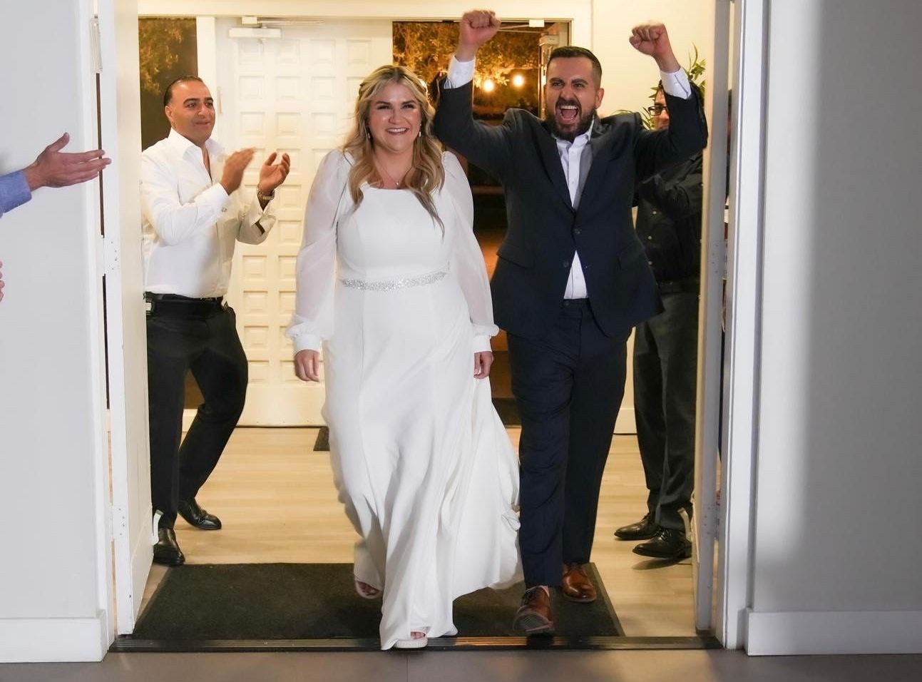 Newlyweds celebrate, exiting a building, man raises arms, people applaud.