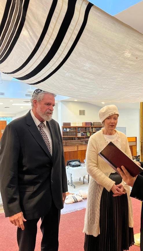 Couple at ceremony under a striped canopy; the man wears a suit and kippah, the woman, a dress and hat.