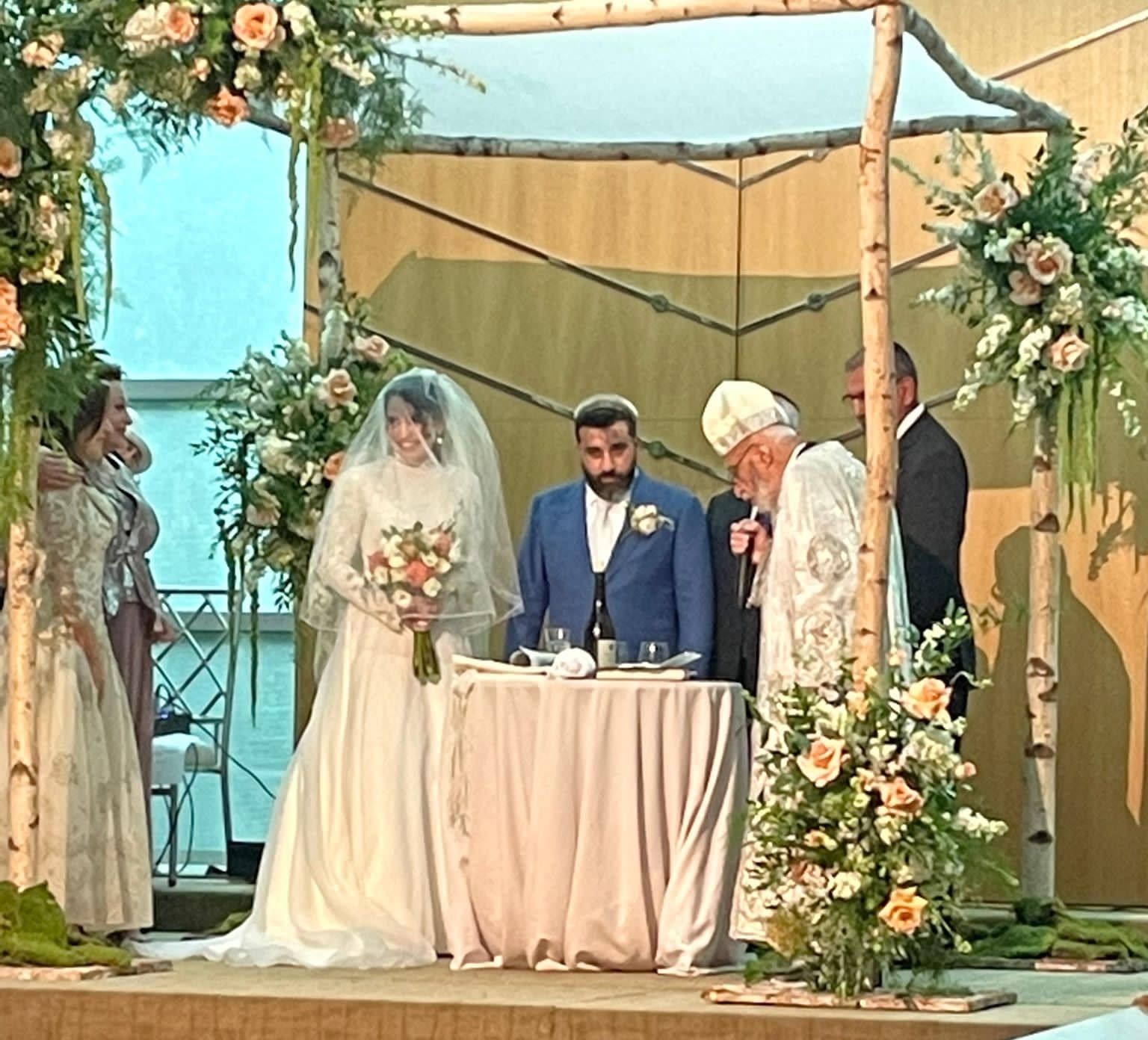Wedding ceremony under a chuppah, bride and groom at altar, officiant, guests.