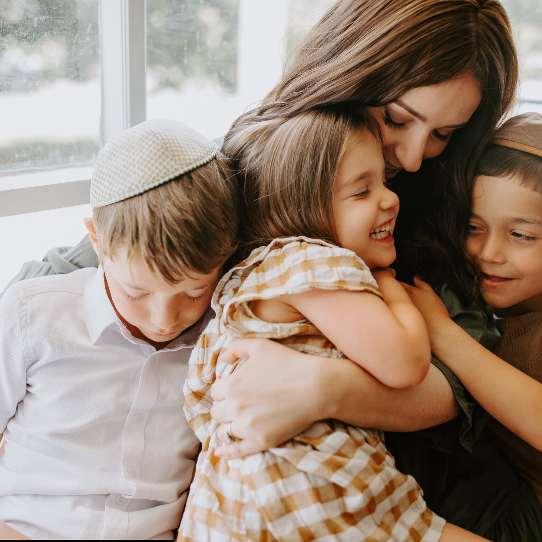 Woman hugging three children, one child wearing a kippah. Natural light, joyful expressions.