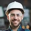 Smiling man in a white hard hat and safety glasses, in a factory setting.