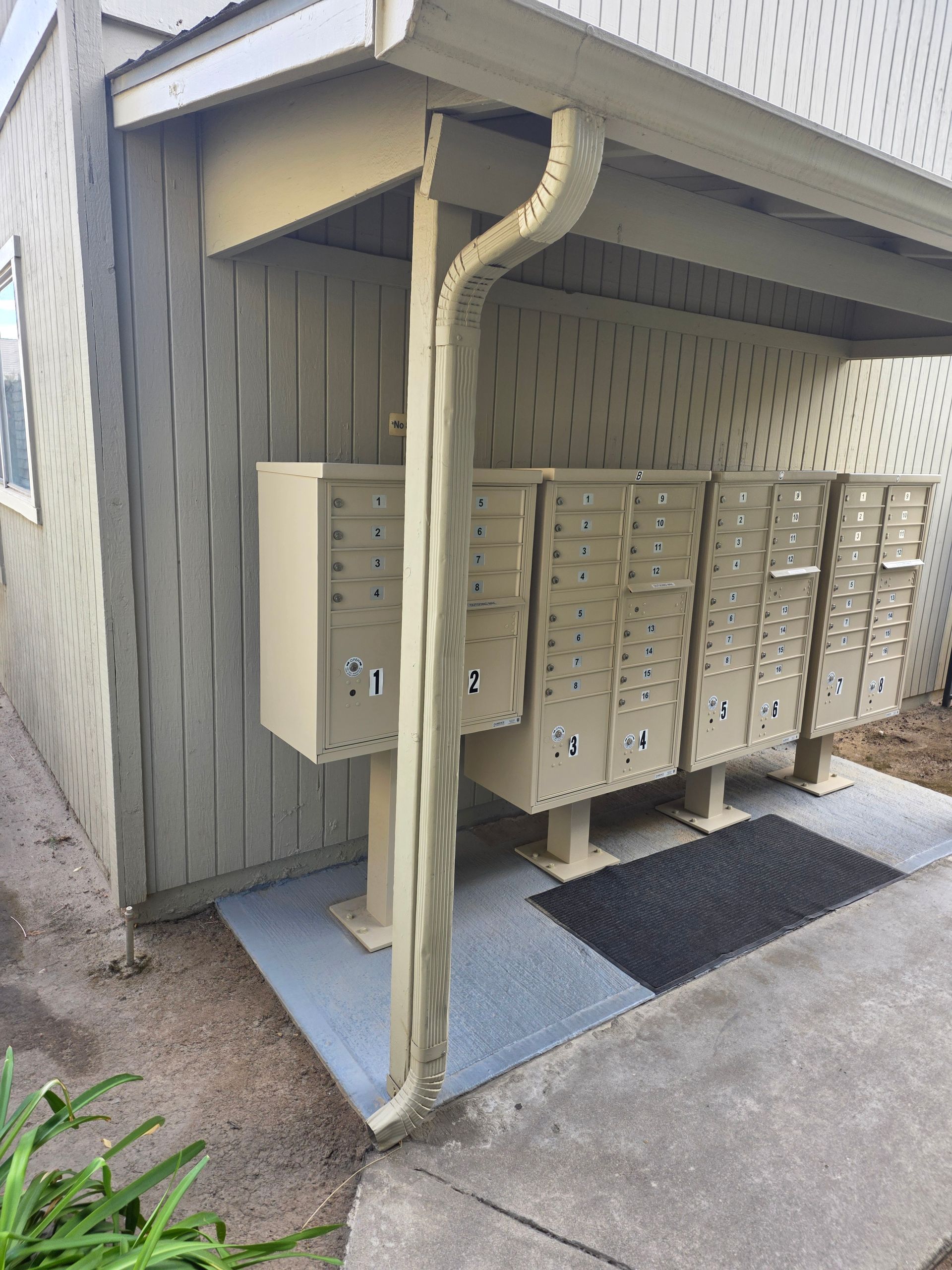 Mailboxes under a covered overhang, next to a building with a gutter. A small ramp and mat are present.