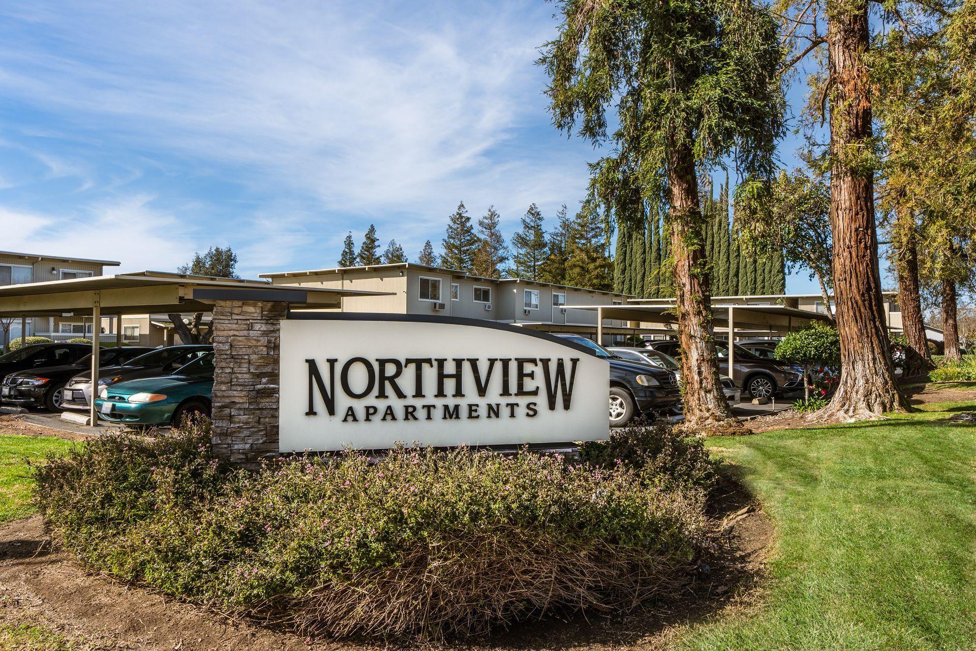 Northview Apartments sign with covered parking and cars. Green grass, blue sky, trees.