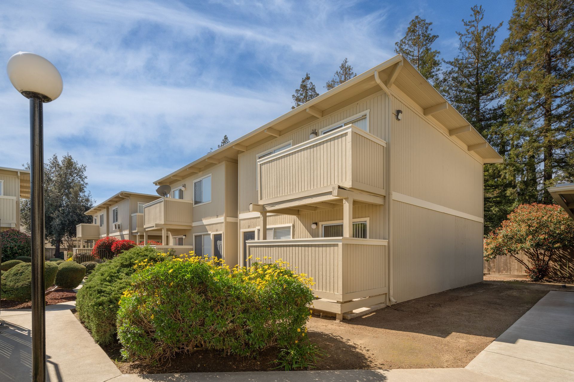 Apartment building with tan siding, balconies, and landscaping on a sunny day.