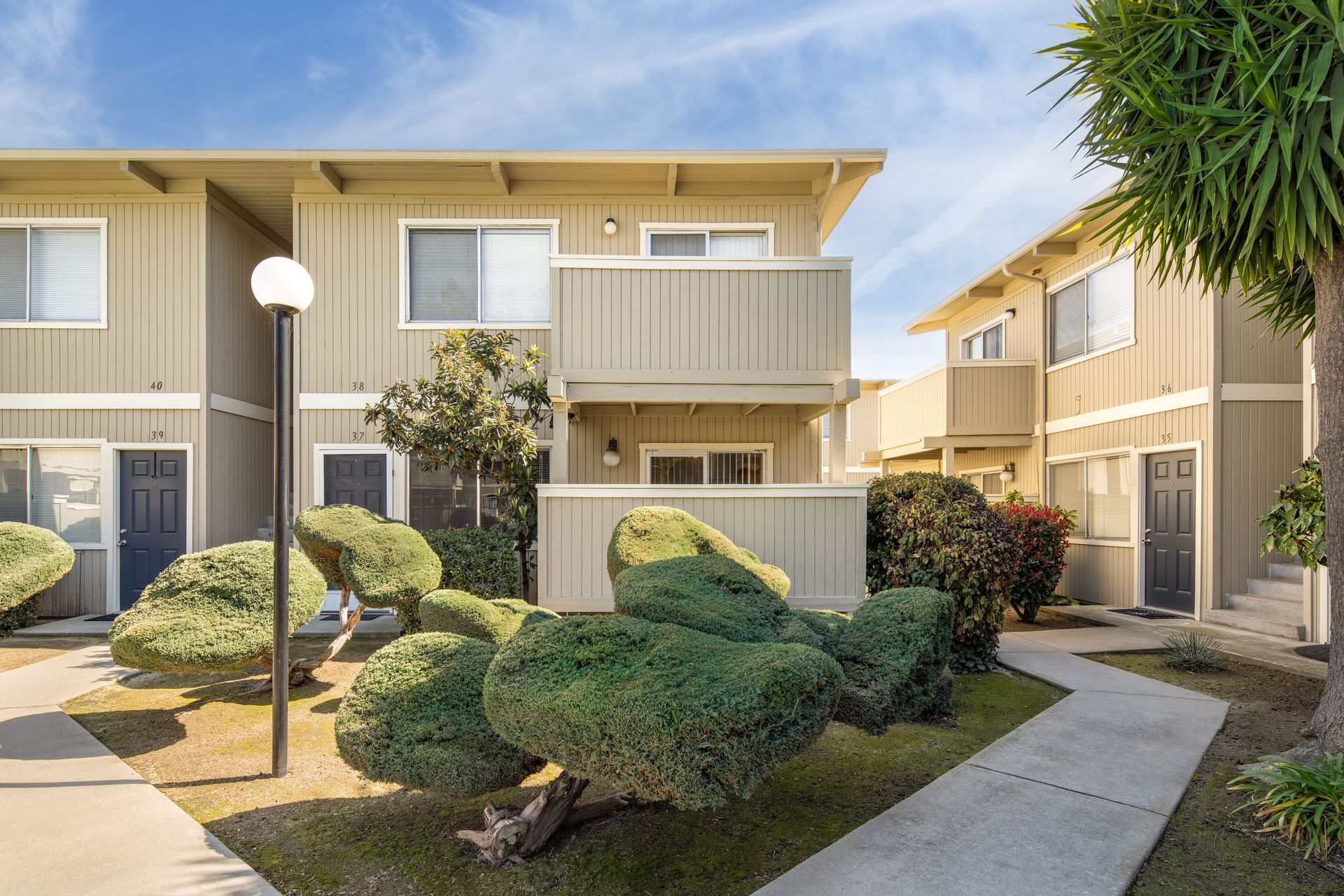 Two-story beige apartment buildings with a walkway, bushes, and a lamppost.