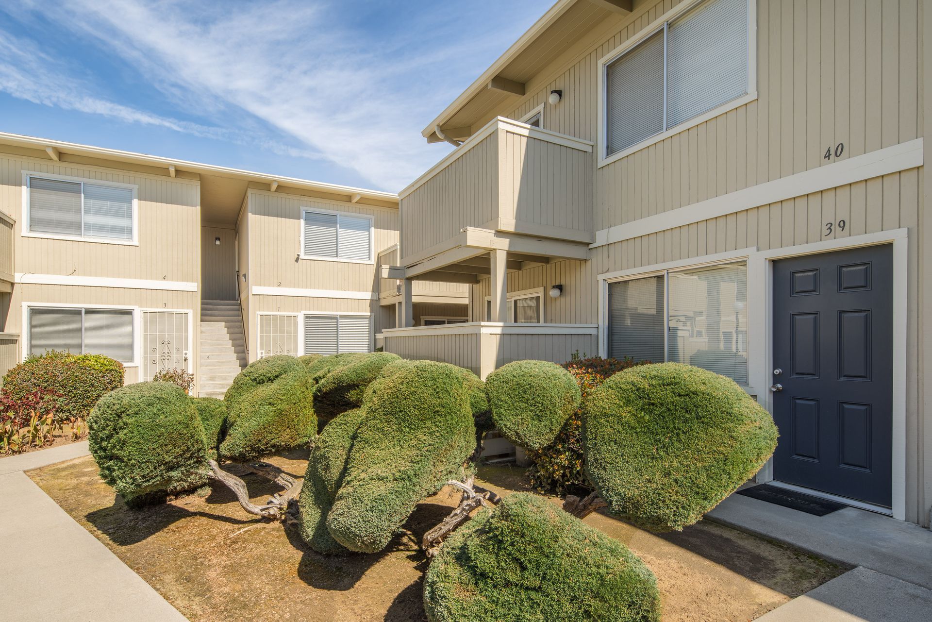 Apartment buildings with light beige siding and blue door, landscaping in front, sunny day.