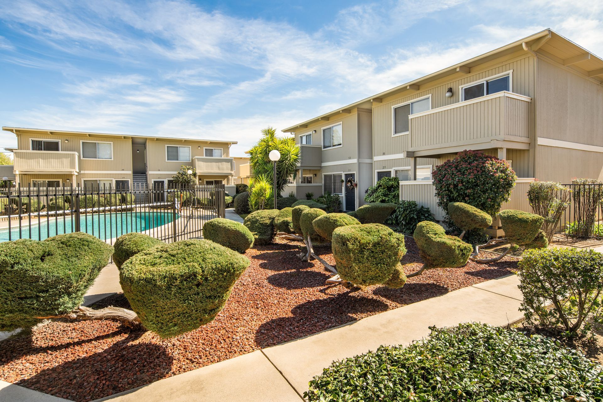 Apartment buildings with balconies and a pool on a sunny day, with bushes and a sidewalk in the foreground.