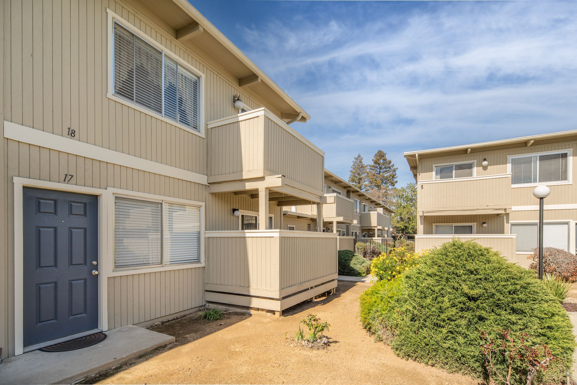 Apartment complex exterior: beige buildings with blue doors and balconies, blue sky and landscaping.