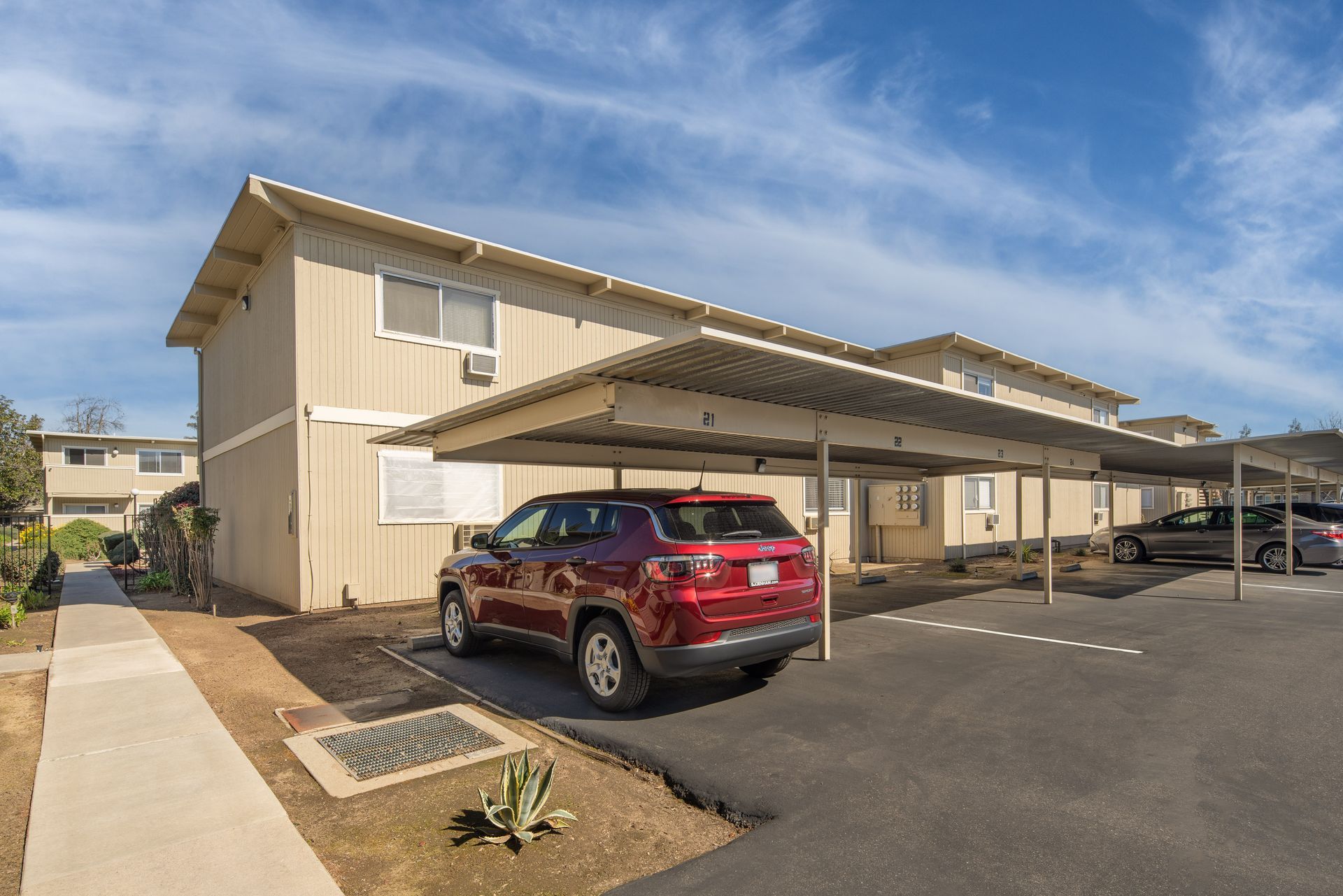 Tan apartment building with carports, red SUV parked under one. Blue sky overhead.