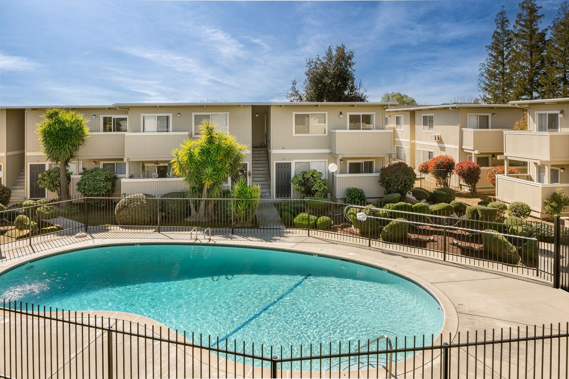 Apartment complex with pool, blue sky. Beige buildings with balconies. Green trees and fence.
