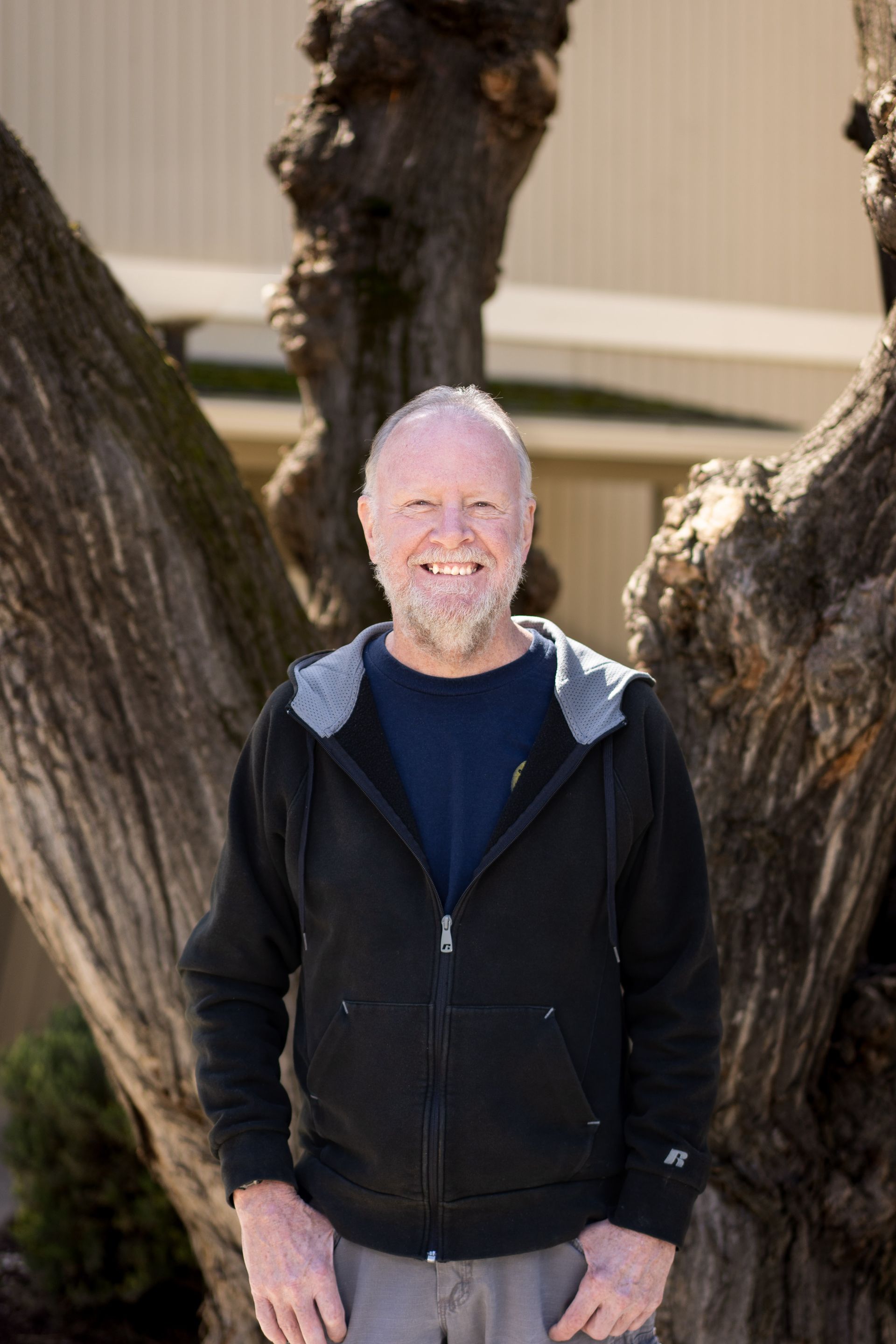 Smiling man with shaved head, gray beard, in black hoodie, standing by tree, outdoors.
