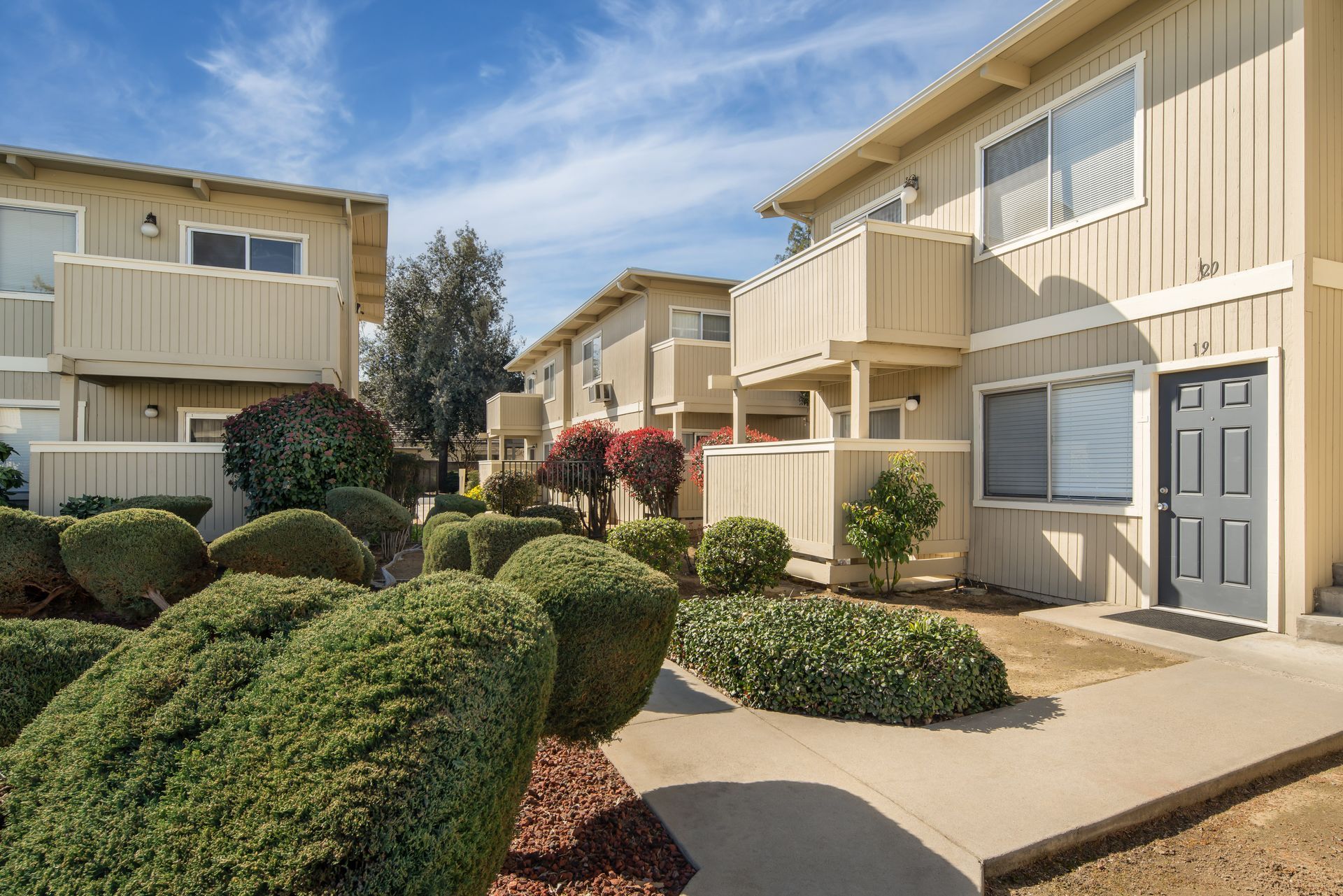 Apartment complex with beige buildings, small balconies, and well-manicured bushes under a blue sky.