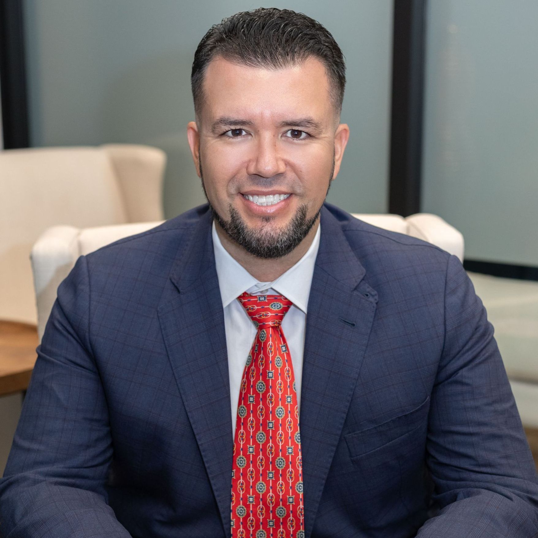 Man in a suit smiles, red tie, seated indoors.