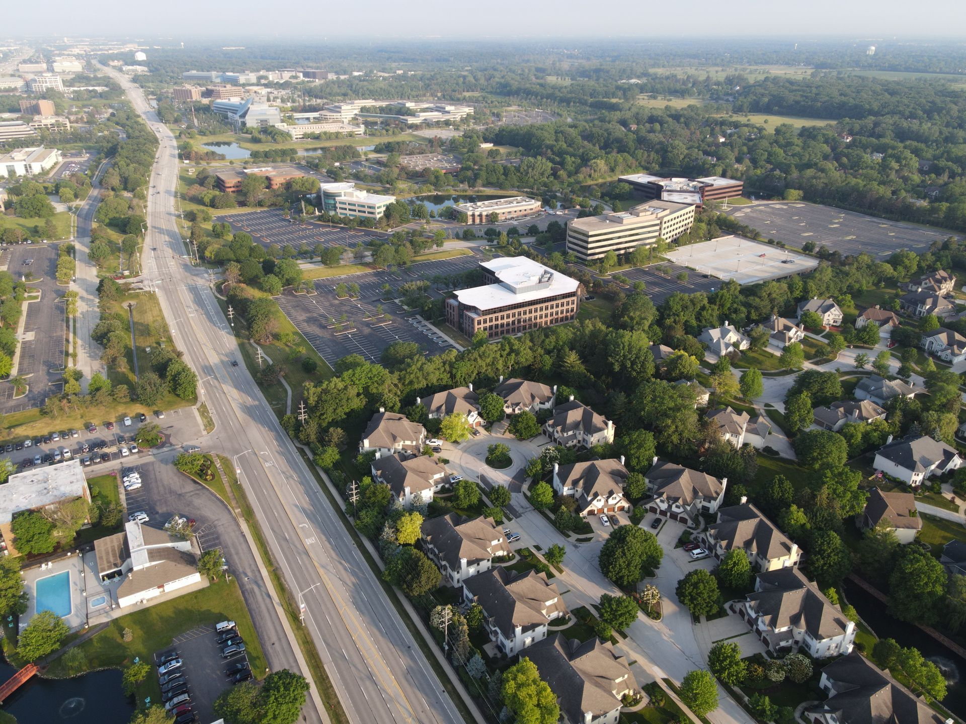 Aerial view of a suburban neighborhood beside a wide road and commercial buildings, with trees and parking lots.