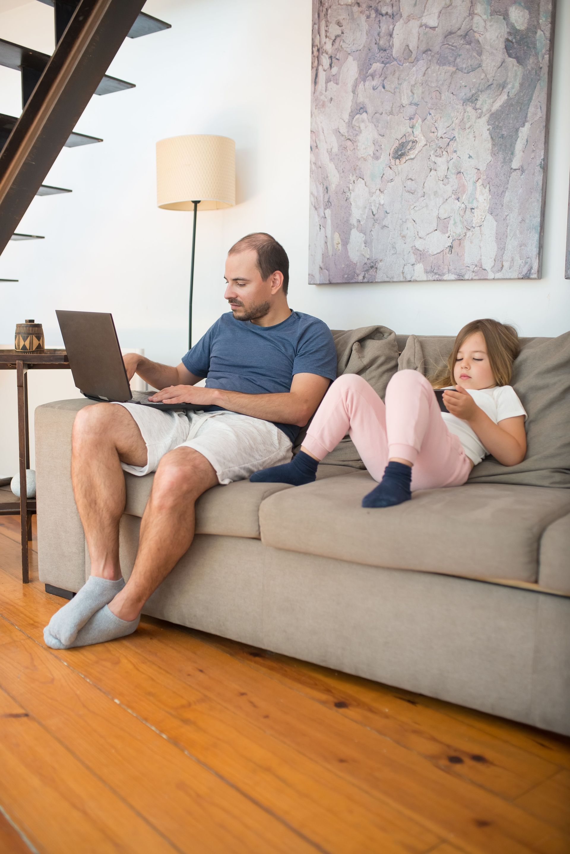 Man using a laptop on a beige couch while a child lies beside him, in a bright living room.