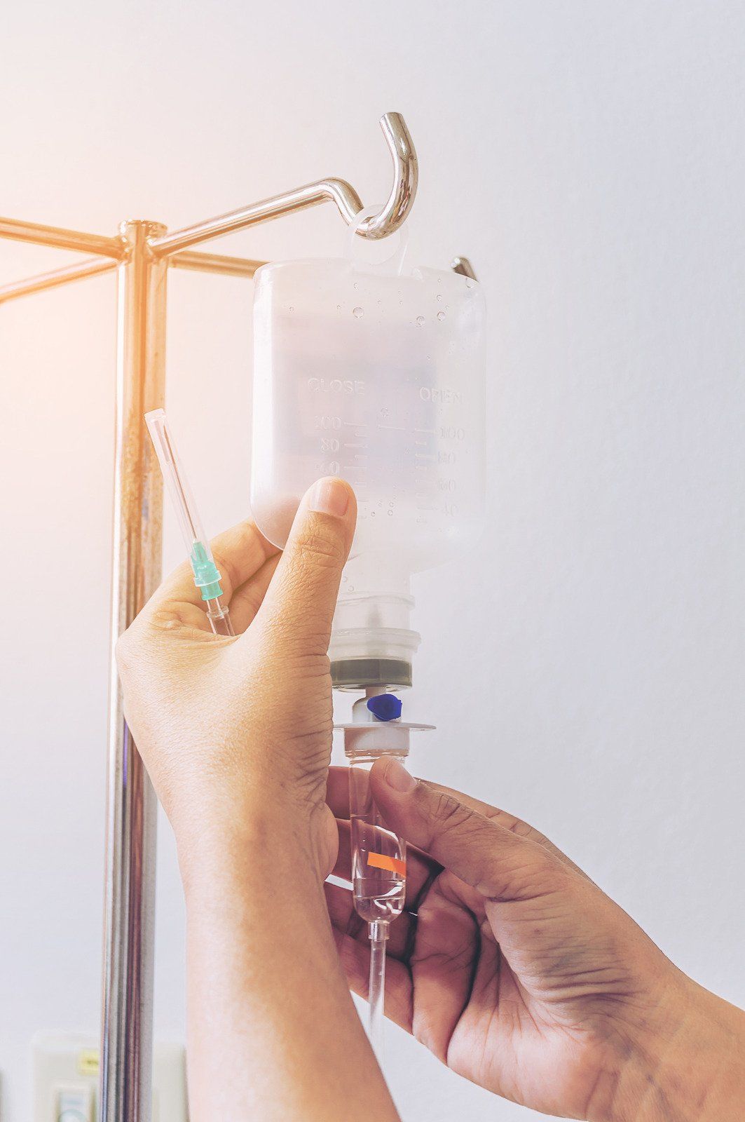 Person's hands adjusting an IV drip bag hanging on a metal stand in a hospital setting.