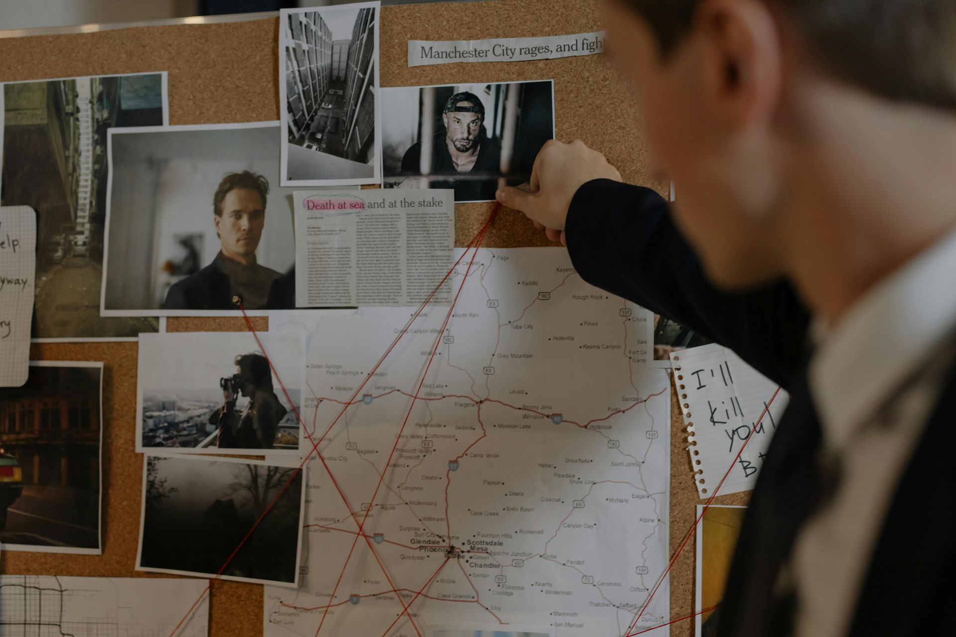 A person examines a corkboard covered in photographs, maps, and red string, typical of a criminal investigation.
