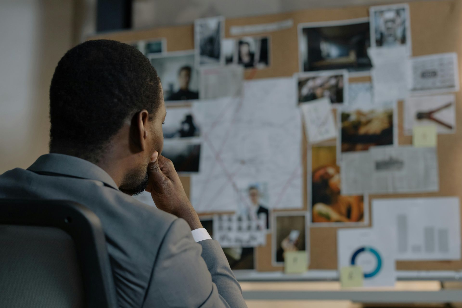 A person in a suit sits with their hand on their chin, studying a corkboard covered with photographs and documents.