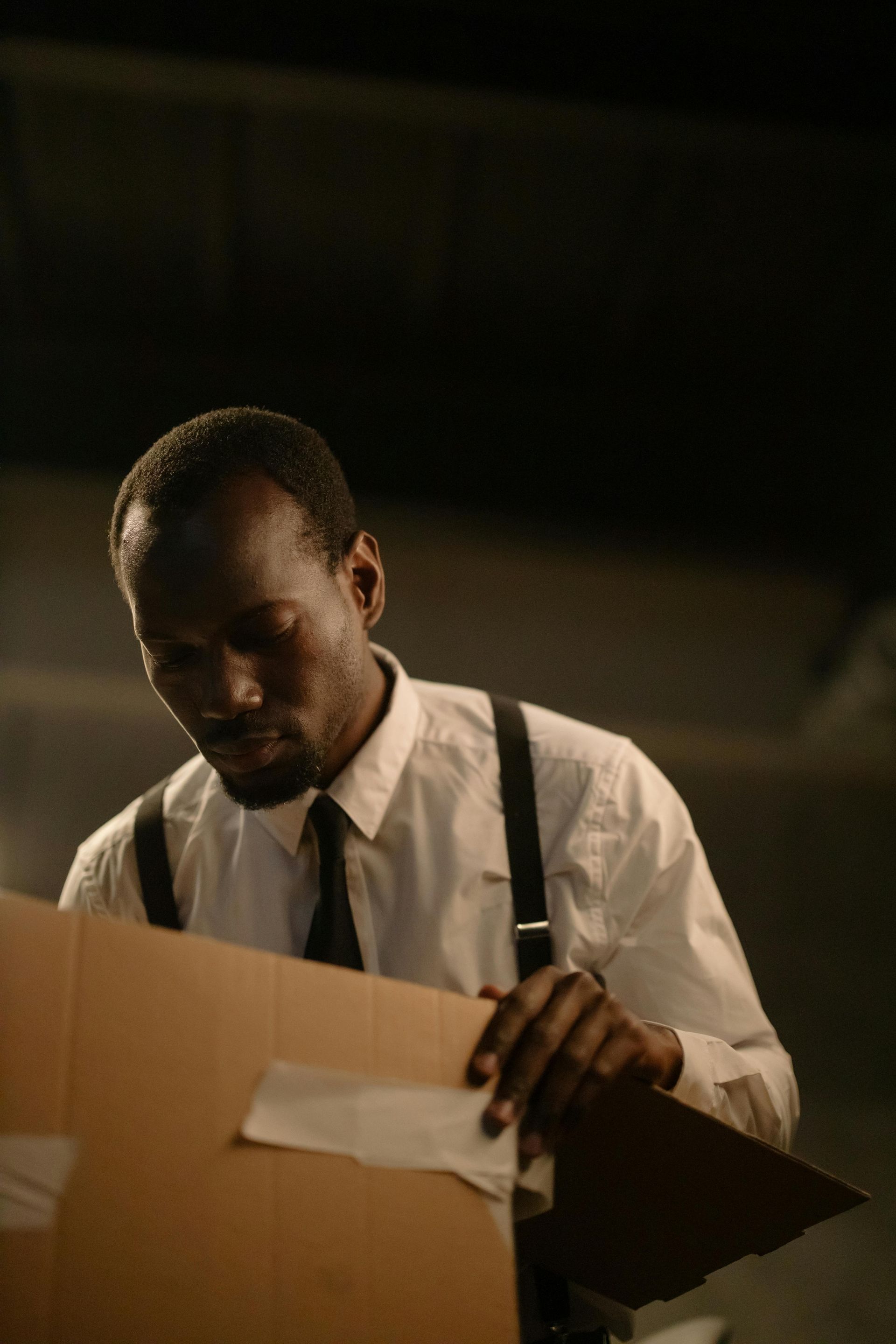 A person in a white shirt and suspenders looks down at a cardboard box while holding a clipboard.