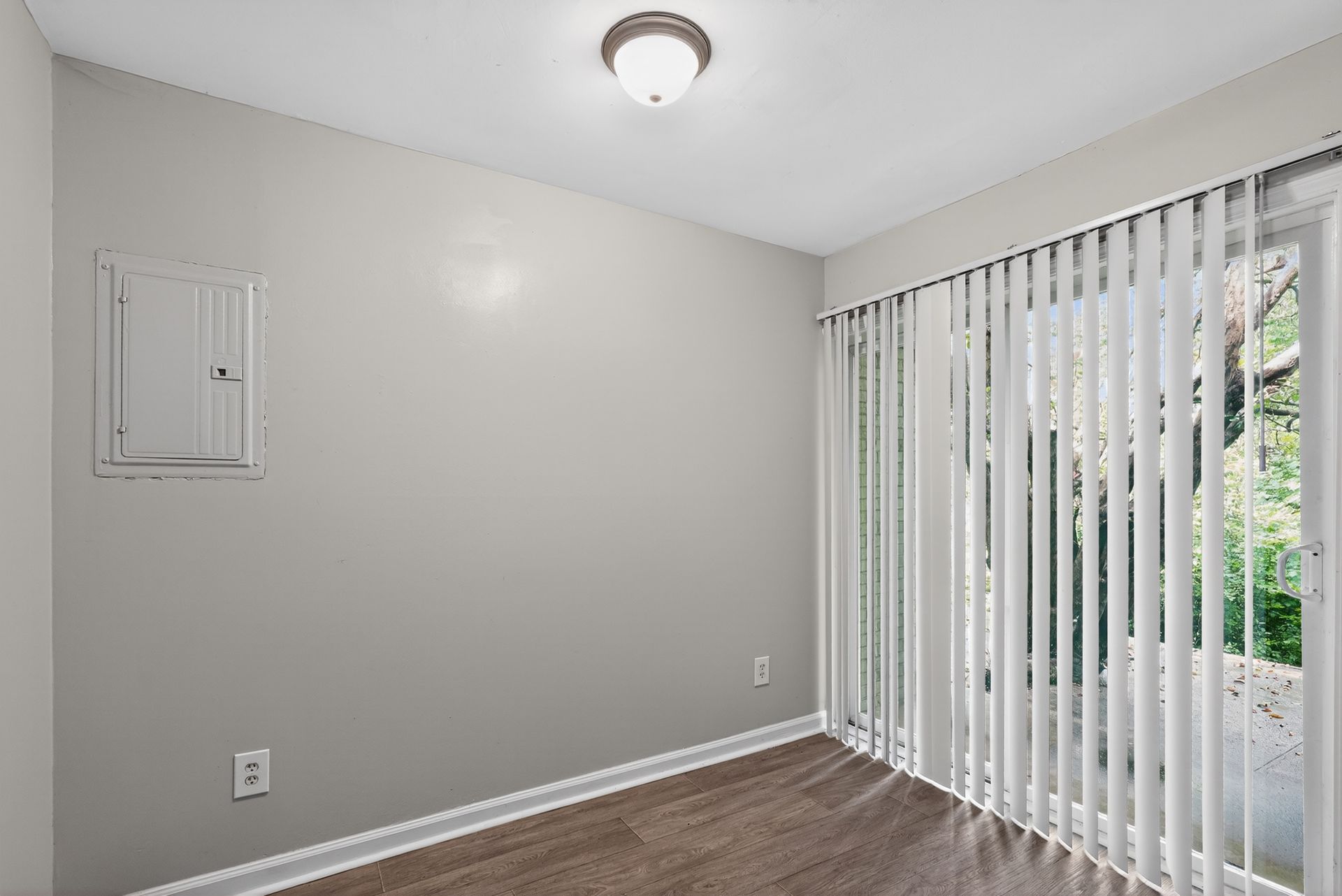 Empty room with gray walls, wood flooring, ceiling light, and sliding glass door with vertical blinds