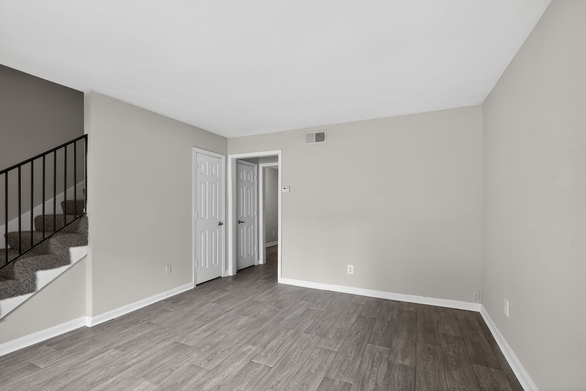 Empty living room with gray walls, wood floor, white door, and staircase railing on the left