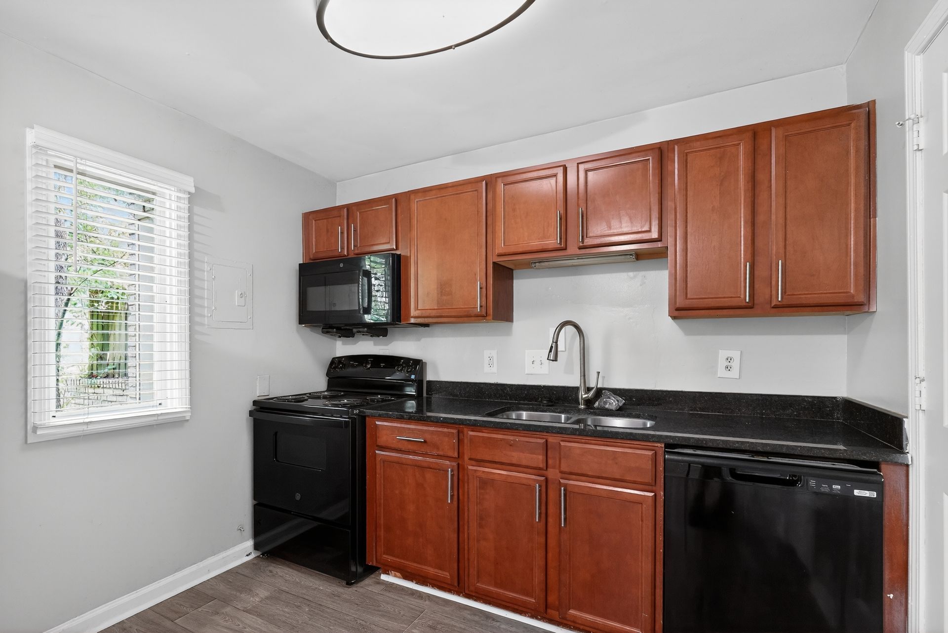 Compact kitchen with cherry cabinets, black appliances, granite counters, and a window with blinds.