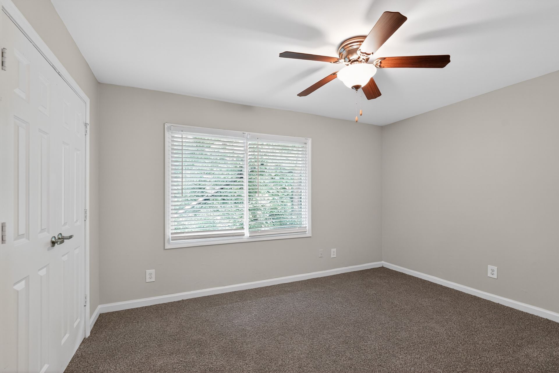 Empty beige bedroom with carpet, white closet doors, window blinds, and a ceiling fan with lights.
