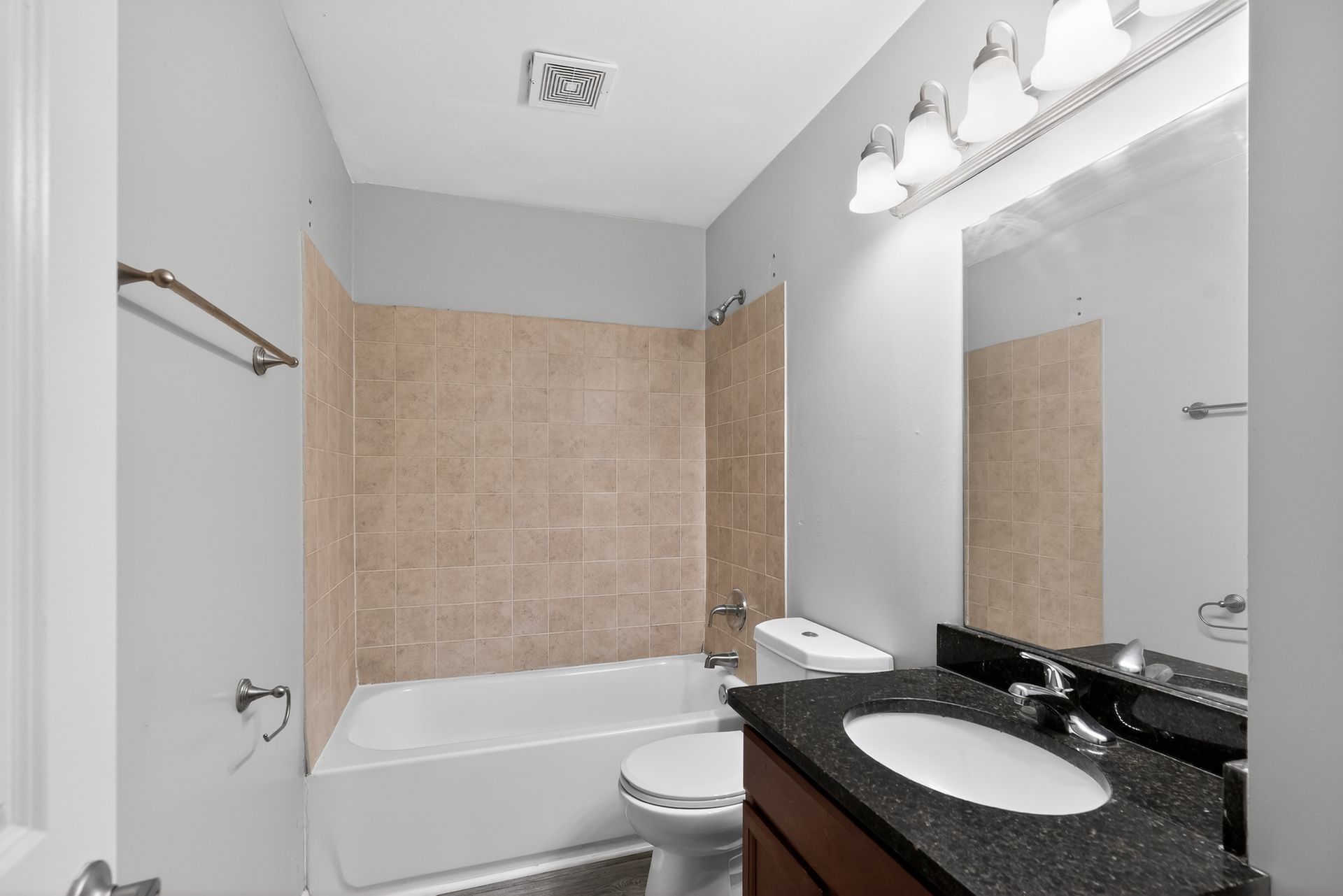 Small beige-tiled bathroom with tub, toilet, and granite vanity under bright lights
