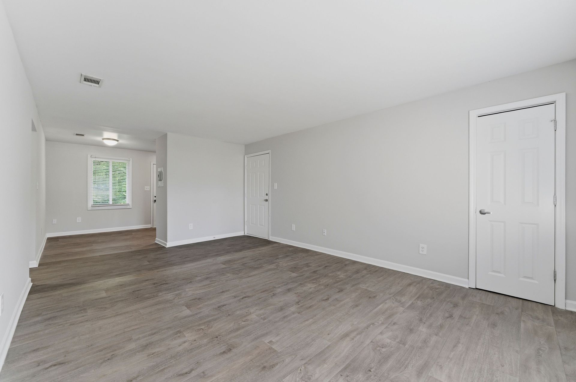 Empty white living room with gray wood flooring, two doors, and a small raised area near a window.