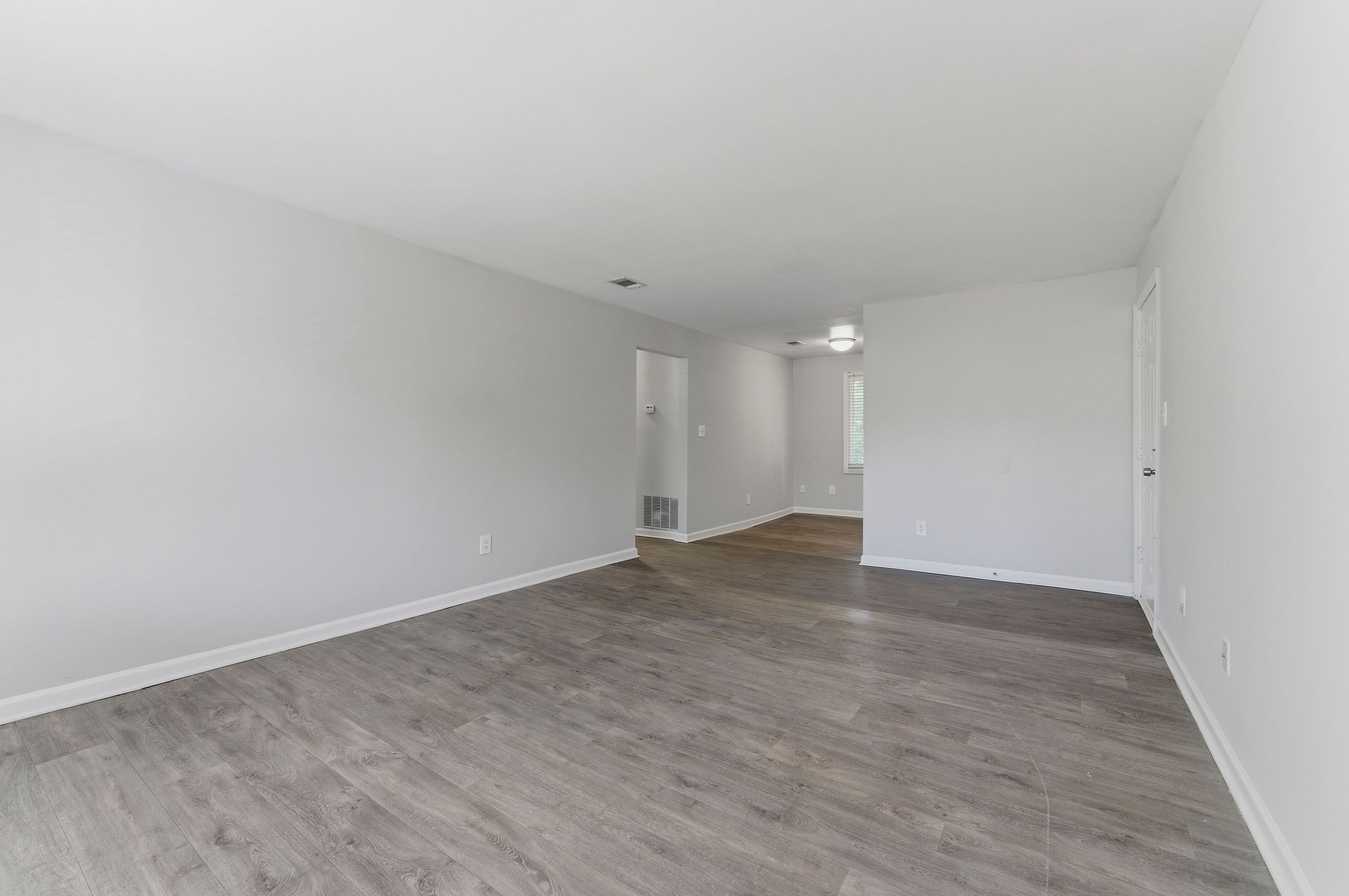 Empty unfurnished room with gray wood flooring and white walls, opening to a hallway in the back.