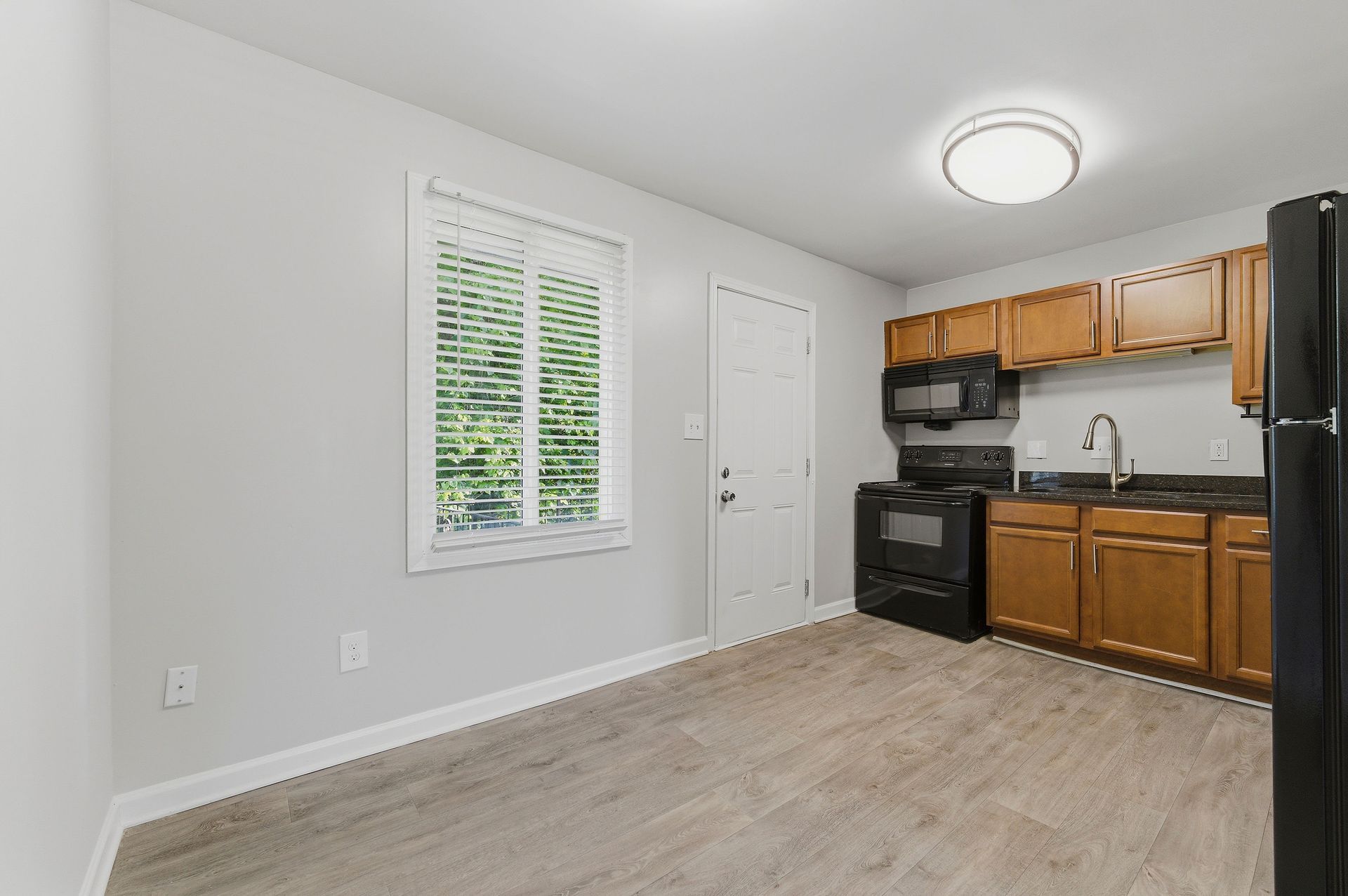 Small kitchen with wood cabinets, black stove, window, white door, and light wood flooring
