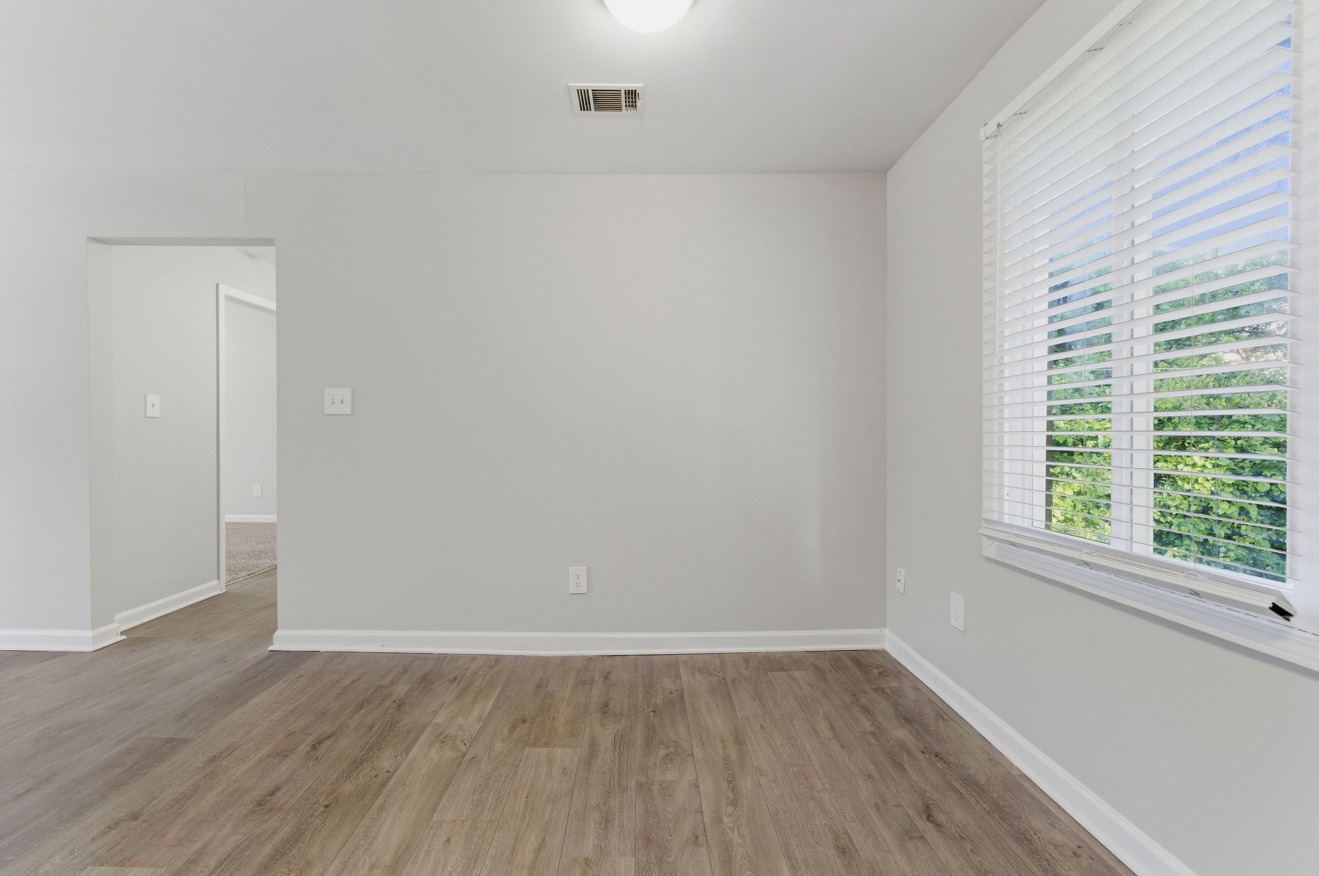Empty white room with wood flooring, doorway on the left, and a window with blinds on the right.
