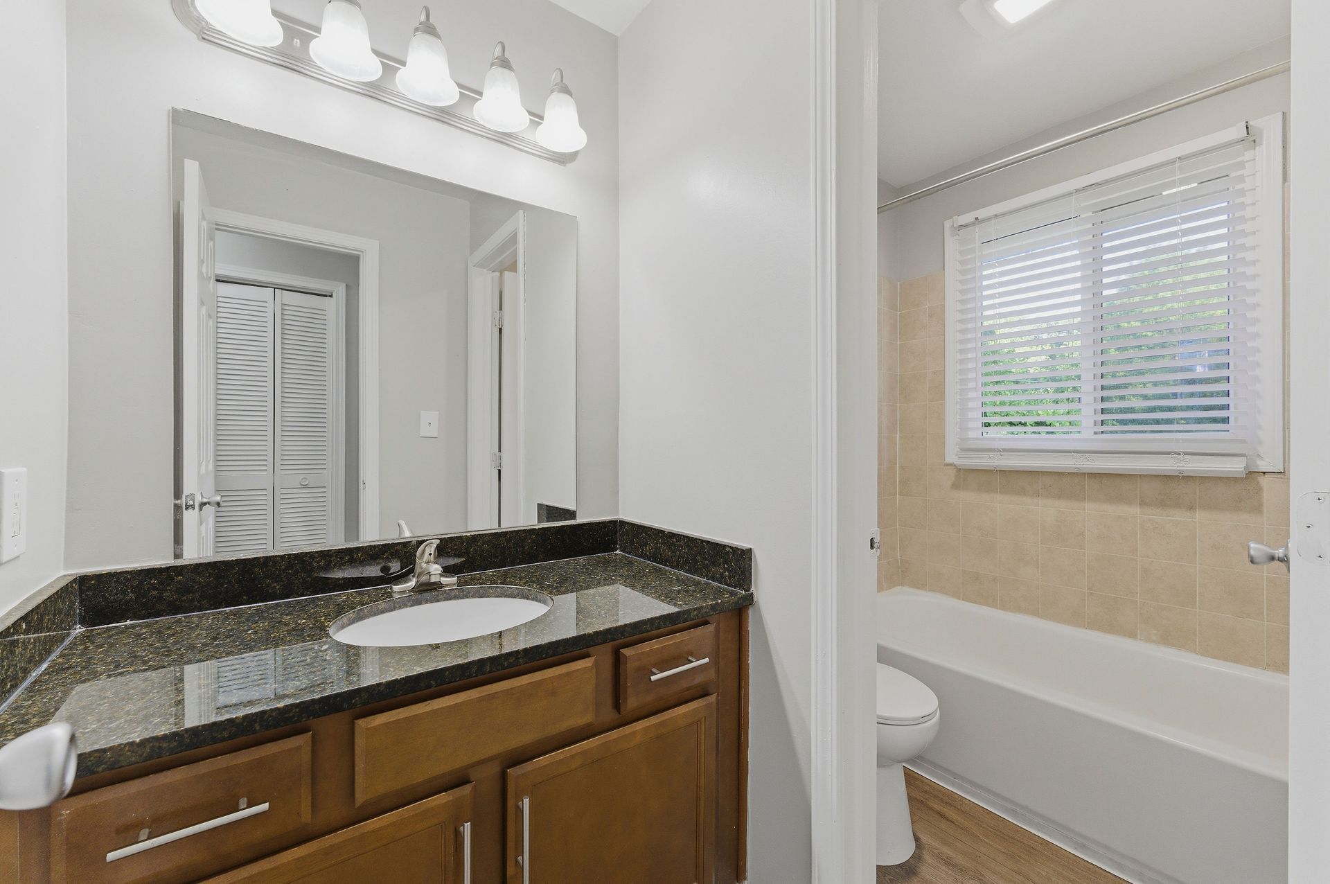 Bathroom with granite vanity, wall mirror, and a bathtub beside a window with blinds.
