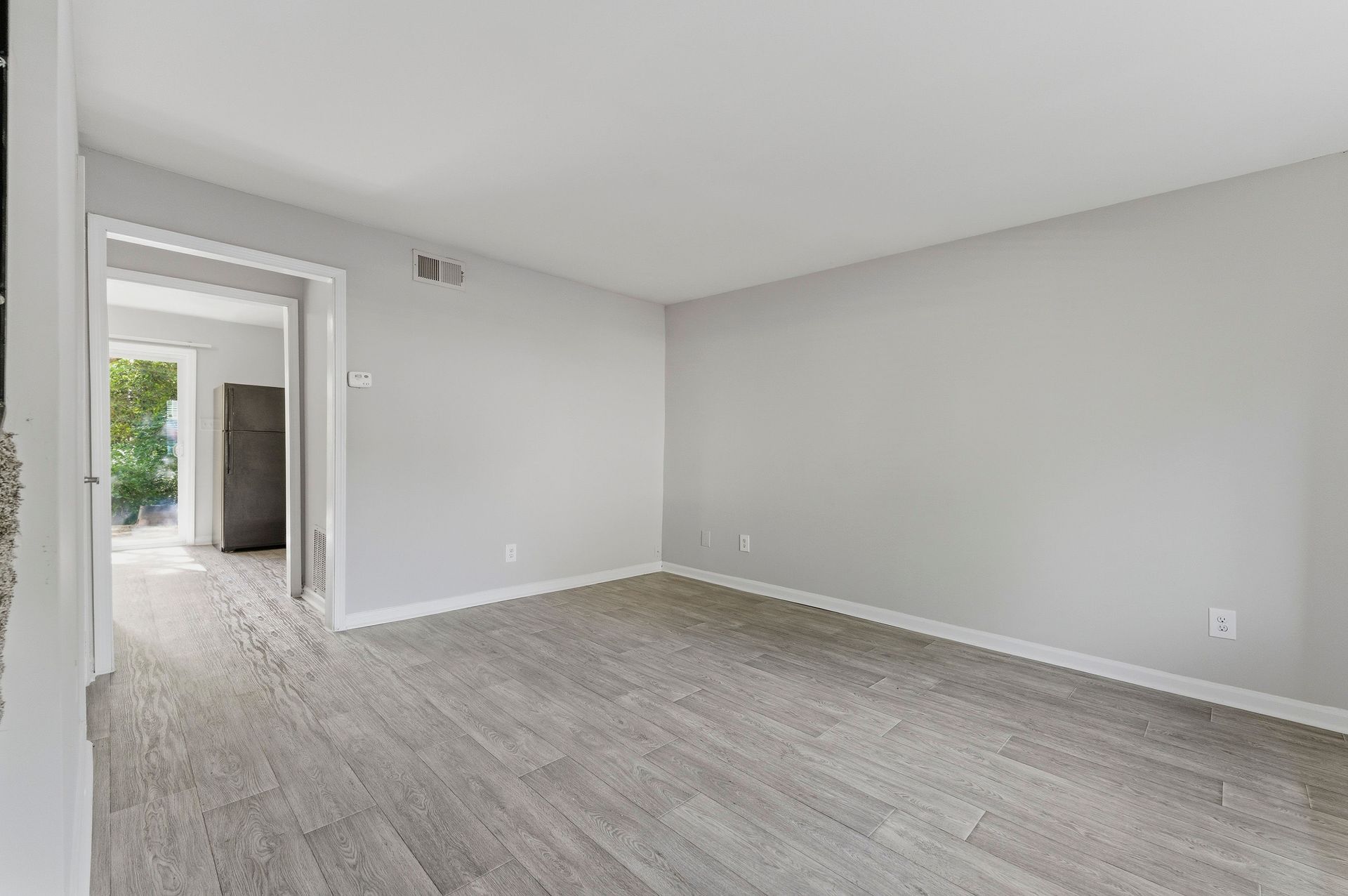 Empty gray living room with carpet, white walls, and a doorway to a brighter hall