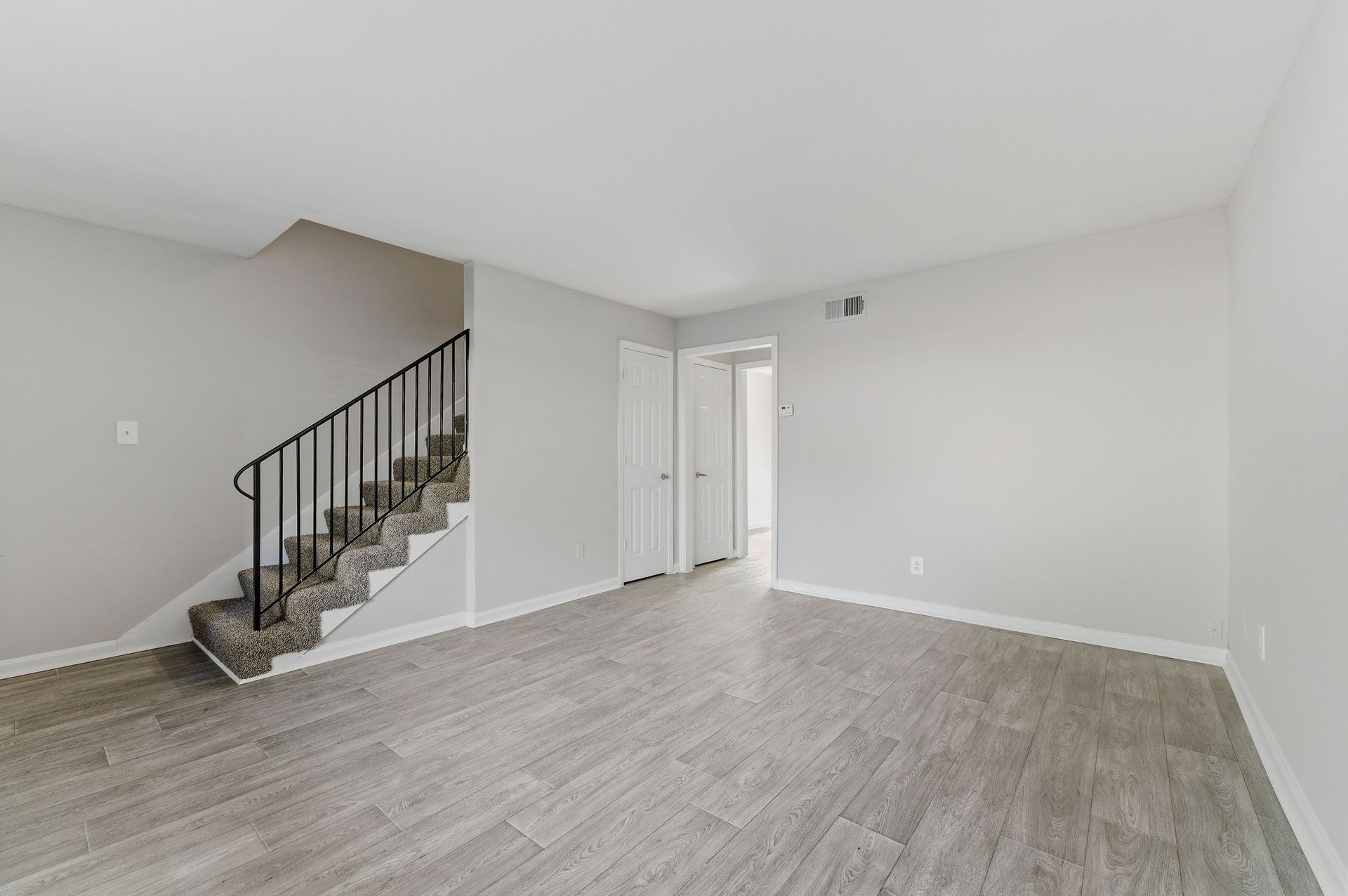 Empty living room with gray flooring, white walls, and carpeted stairs with black railing