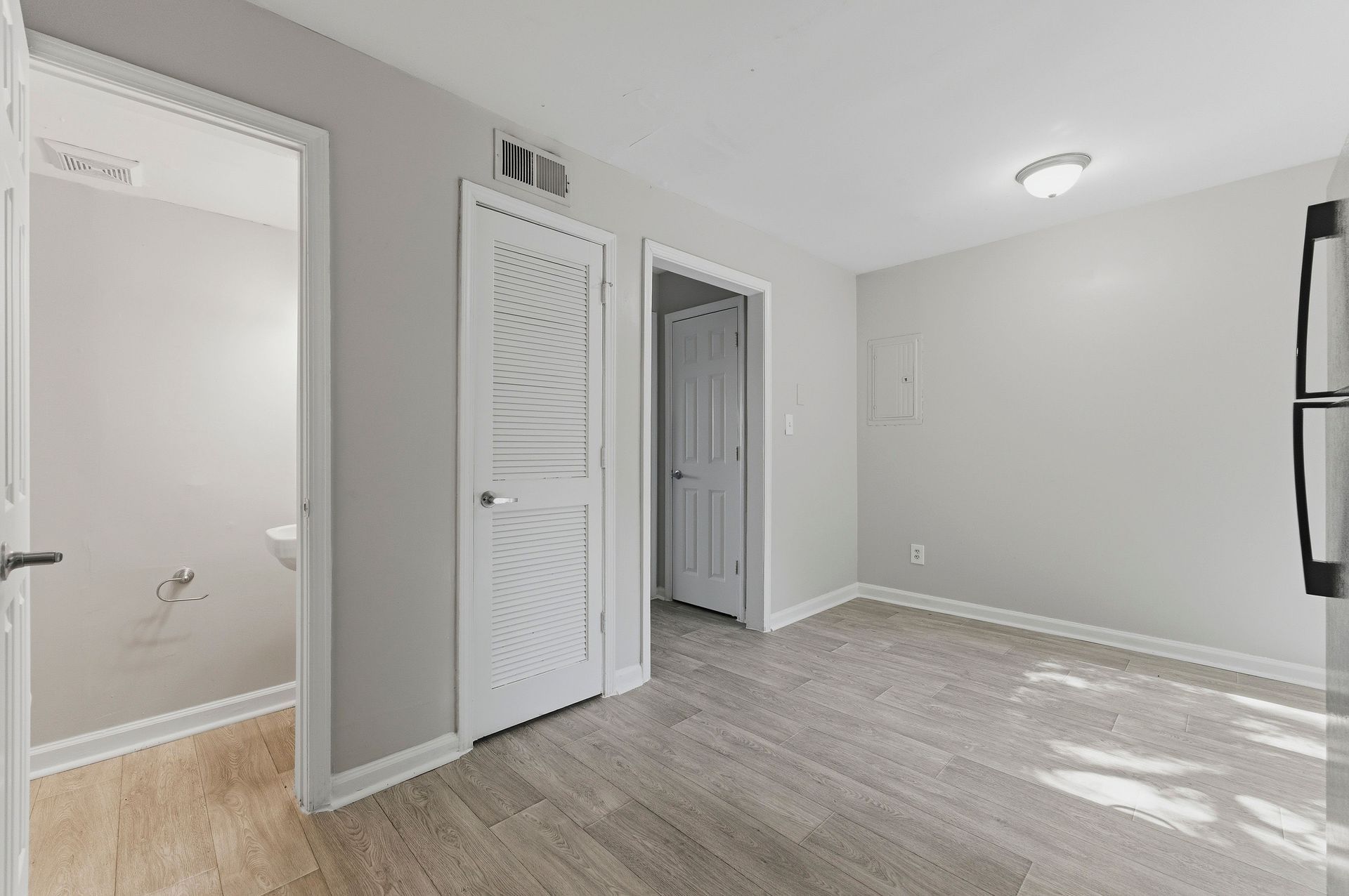 Empty white room with light wood flooring, closet doors, and a doorway to a bathroom.