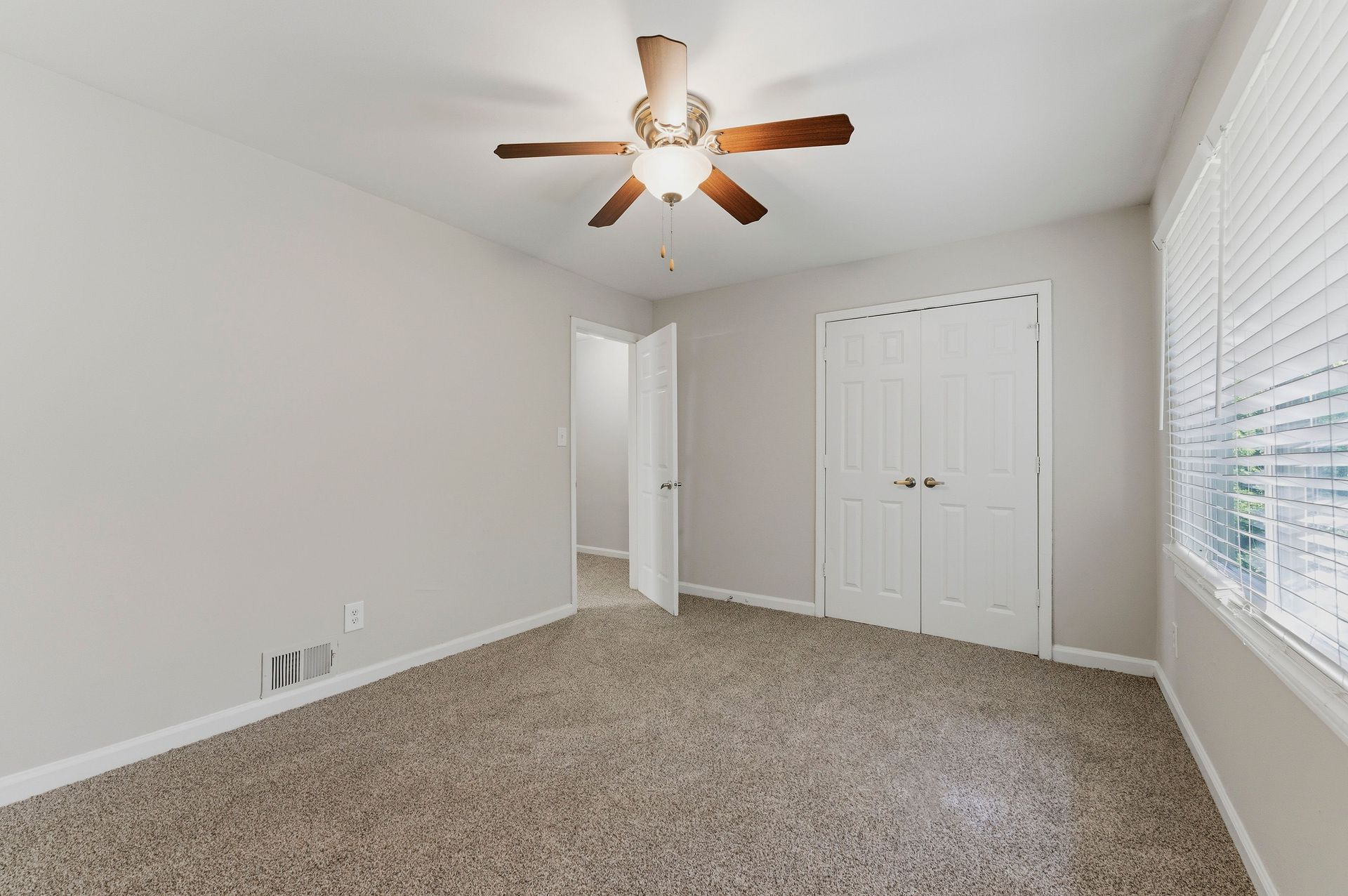 Empty carpeted bedroom with white walls, ceiling fan, closet doors, and window blinds