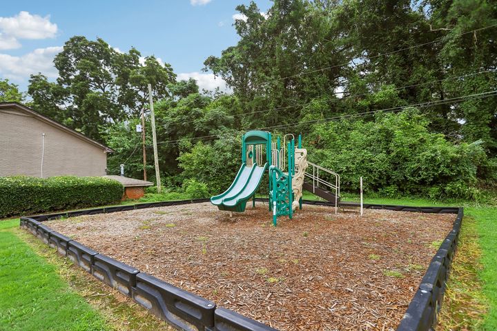 Empty playground with a green slide in a leafy park, bordered by a low fence and wood chips.