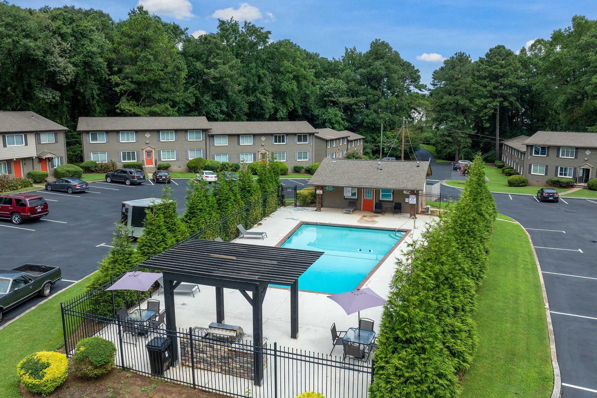 Apartment complex pool with turquoise water, pergola seating, and parked cars on a sunny day