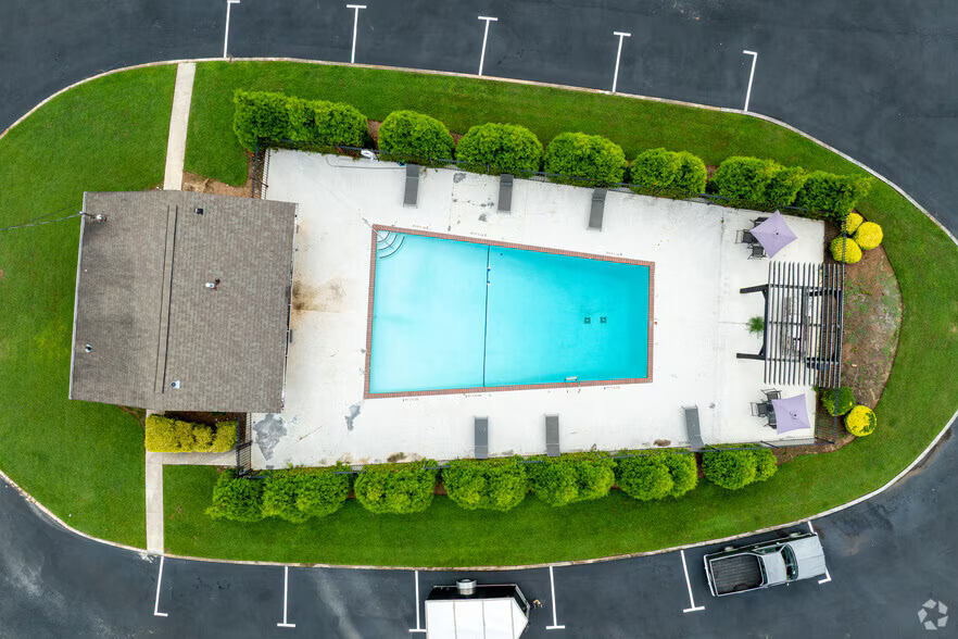 Aerial view of a backyard pool with patio, loungers, and a surrounding hedge, next to a house and parking lot