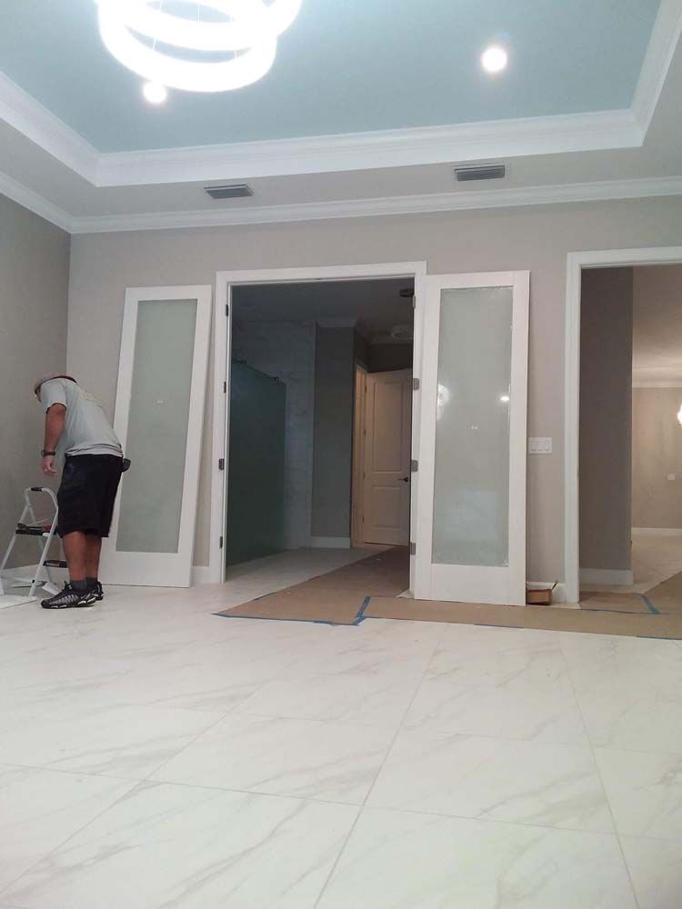 Man on ladder painting around doorway in a room with light grey walls and marble-like floors.