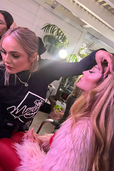 Una maquilladora maquilla a una mujer con un abrigo de piel rojo, en un entorno interior luminoso.