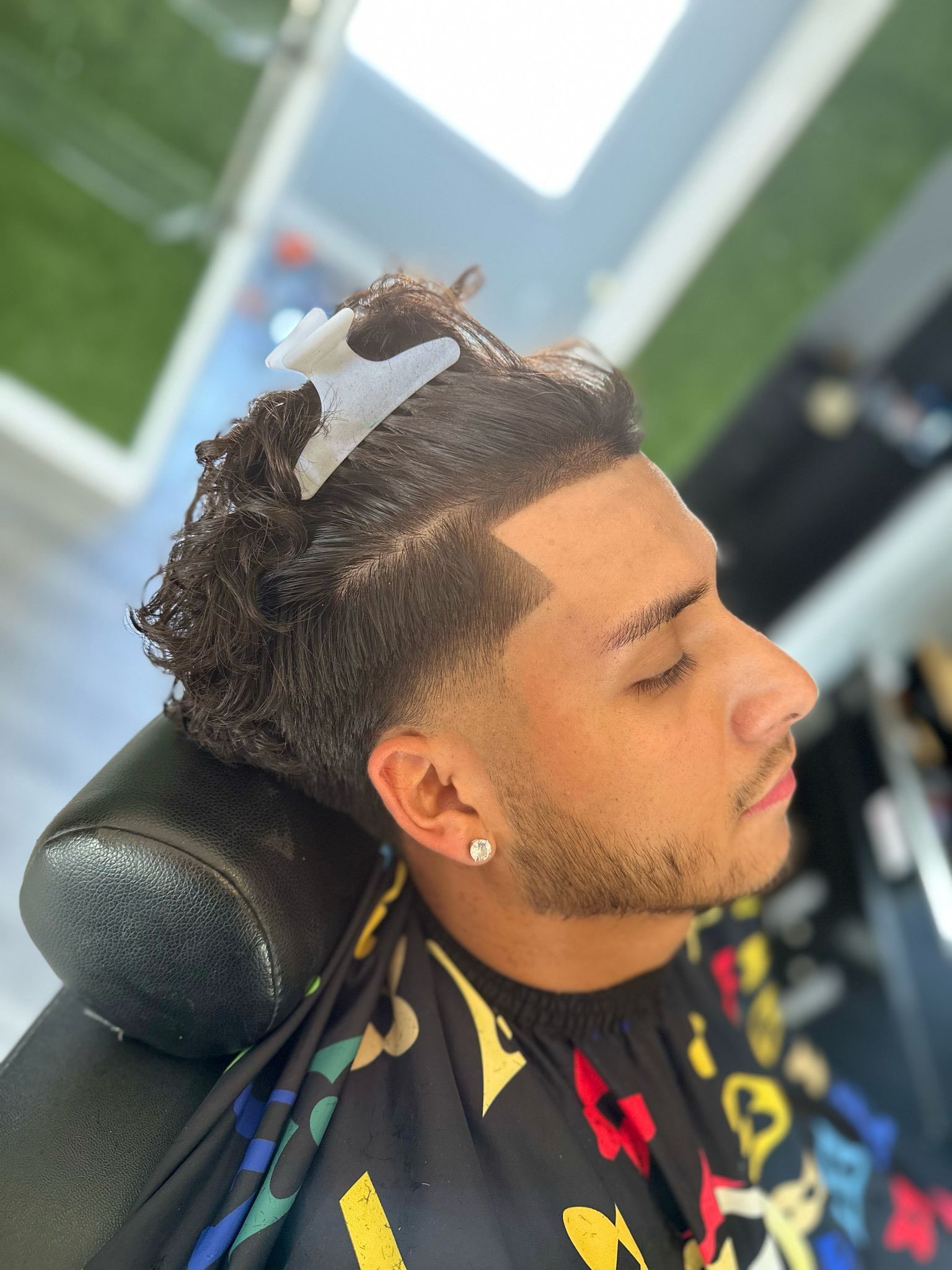 Hombre de pelo rizado en una barbería, cortándose el pelo. Lleva una rueda dentada, una pinza para el pelo y una capa estampada.