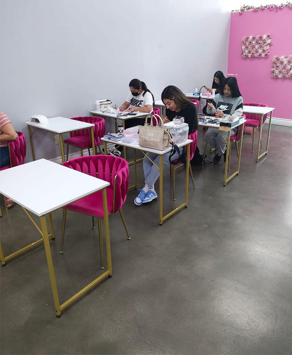 Nail art class: Students at individual desks with pink chairs, working on nails in a bright pink and white room.