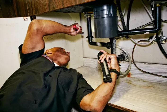 A person in a black shirt works under a kitchen sink, using a flashlight to inspect garbage disposal plumbing pipes.