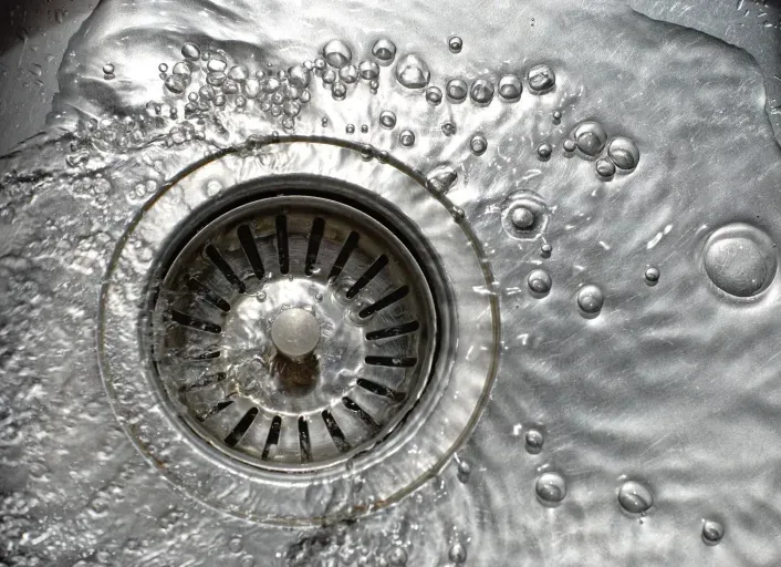 Water flows from a faucet into a metal kitchen sink drain, creating bubbles on the surface.